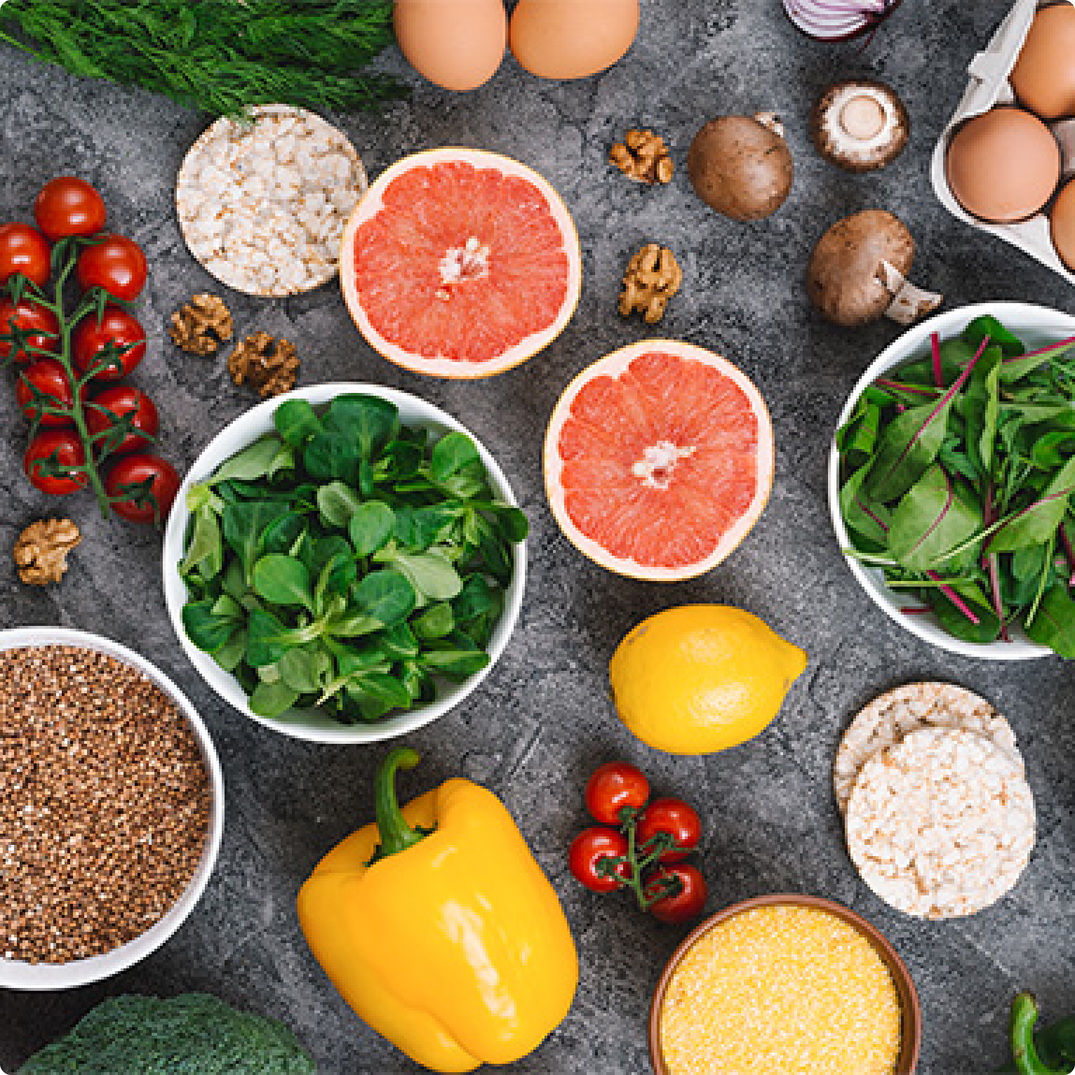Flat lay of fresh fruits, vegetables, grains, and herbs including grapefruit, spinach, tomatoes, lemon, oats, and mushrooms on a dark surface.